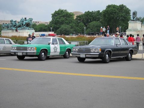 1991 Ford LTD Crown Victoria - green-and-white Sheriff patrol car and a dark four-door sedan parked near a bronze statue and crowds on the Mall.