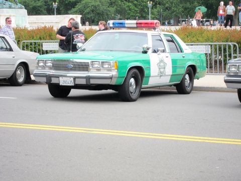 1991 Ford LTD Crown Victoria - green-and-white Sheriff patrol car with a Ford grille and roof lightbar moving along a street past onlookers.