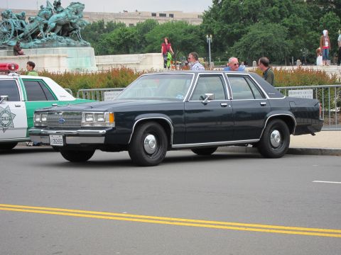 1991 Ford LTD Crown Victoria - dark blue Ford four-door sedan parked at the curb near a bronze equestrian statue with spectators behind it.