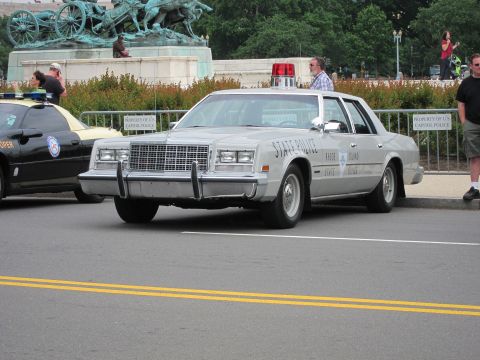 1991 Ford LTD Crown Victoria - silver Rhode Island State Police sedan with a push bar and roof light rolling along a street past a bronze statue.