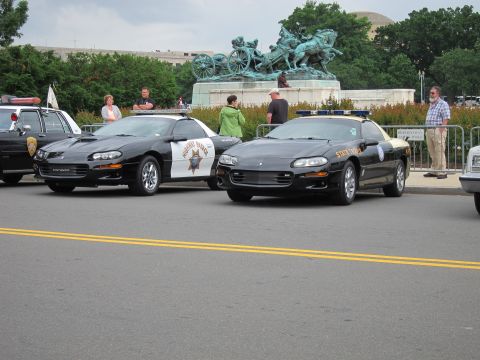 1991 Ford LTD Crown Victoria - Two black Chevrolet Camaro patrol cars, one a Highway Patrol unit, parked near a bronze statue with onlookers behind.