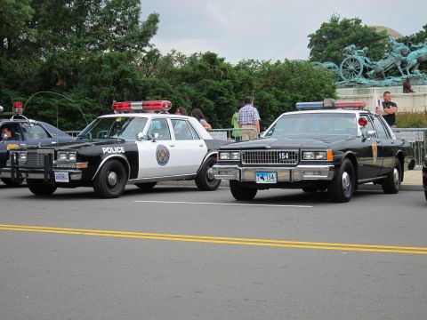 1991 Ford LTD Crown Victoria - black-and-white Police sedan parked beside a dark patrol car numbered 154 near a bronze statue and spectators.