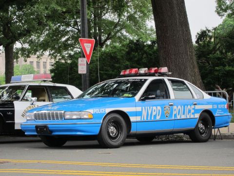 1991 Ford LTD Crown Victoria - light blue NYPD police sedan with white striping parked beside a yield sign, another cruiser to its left.