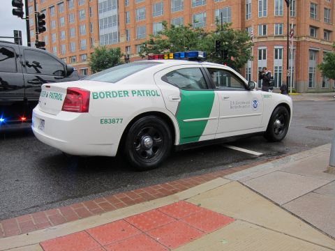 1991 Ford LTD Crown Victoria - white-and-green US Border Patrol sedan with a roof light bar parked along a curb near brick buildings, rear view.