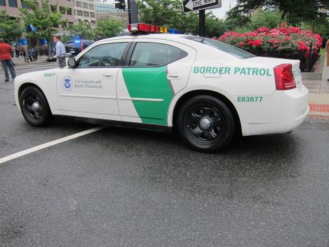 1991 Ford LTD Crown Victoria - white sedan in U.S. Customs and Border Protection Border Patrol livery, numbered E83877, parked on a rainy urban street.