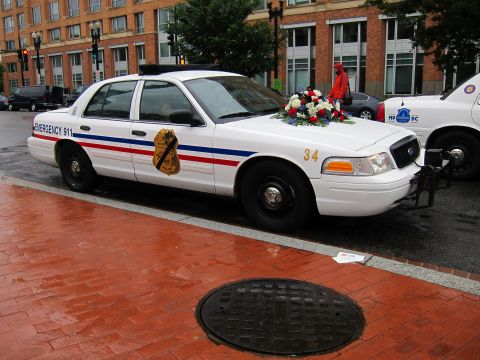 1991 Ford LTD Crown Victoria - white Emergency 911 police sedan numbered 34 sits on wet brick pavement with a memorial floral wreath placed on its trunk.