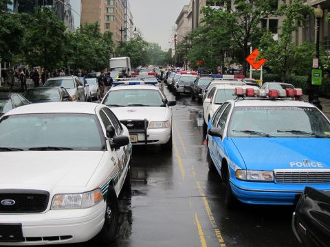 1991 Ford LTD Crown Victoria - long row of police cars, including white and bright-blue memorial sedans, parked nose-to-tail along a wet downtown street.