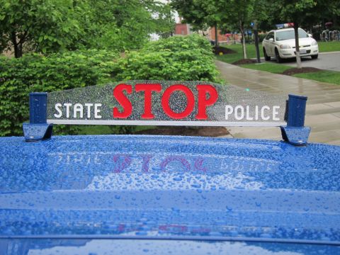 1991 Ford LTD Crown Victoria - chrome STATE POLICE STOP hood sign with red lettering on a rain-beaded blue hood, with shrubs and a white car behind.