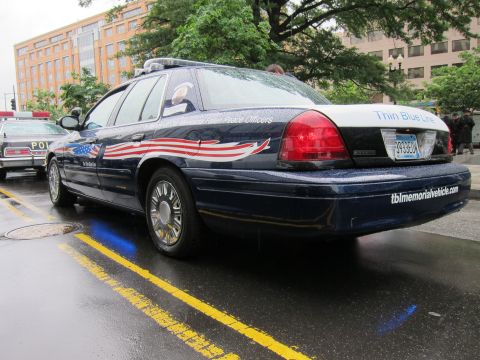 1991 Ford LTD Crown Victoria - Rear quarter of a dark blue Thin Blue Line memorial police sedan with stars-and-stripes graphics, parked on a wet street.
