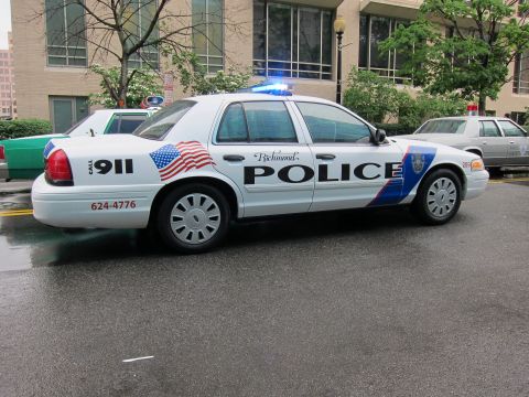 1991 Ford LTD Crown Victoria - Rear three-quarter view of a white Richmond Police sedan with flag and blue swoosh graphics on a rain-slicked street.