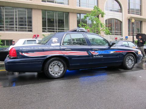 1991 Ford LTD Crown Victoria - dark blue Thin Blue Line memorial police sedan with flag graphics reading 'In Memory Of Our Minnesota Fallen Peace Officers' on a wet stree…