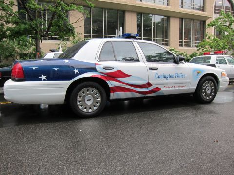 1991 Ford LTD Crown Victoria - white Covington Police sedan wrapped in American flag graphics with the slogan 'United We Stand' parked on a wet street.
