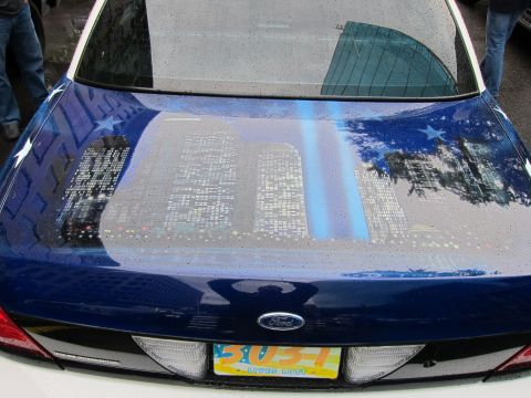 1991 Ford LTD Crown Victoria - Rear trunk of a blue Ford painted with a glowing nighttime city skyline and twin light beams, above a yellow license plate.