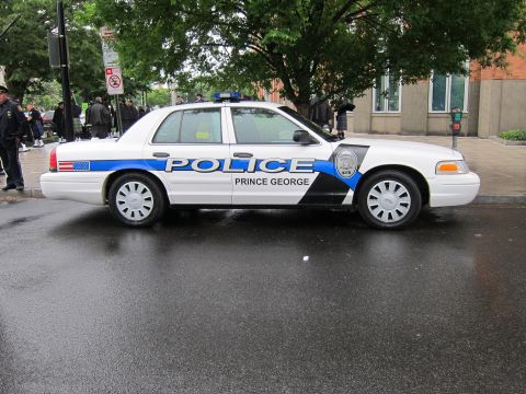 1991 Ford LTD Crown Victoria - Side profile of a white Ford Crown Victoria police car marked POLICE Prince George with a blue accent stripe on a wet street.