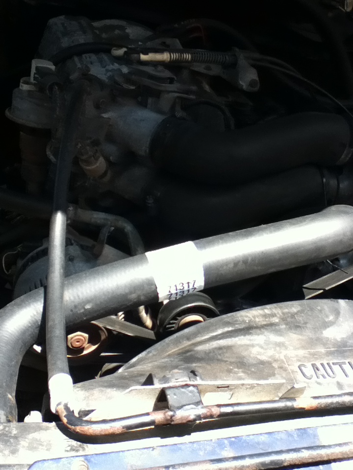 Engine bay with hoses, belts, and fan visible. A white label with numbers is on a hose