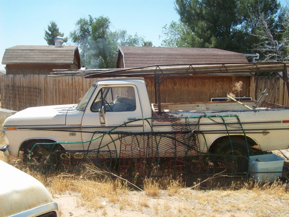Side view of a white pickup truck with a custom metal rack over the bed. The truck appears to be from the 1970s and is parked in a dry, grassy area