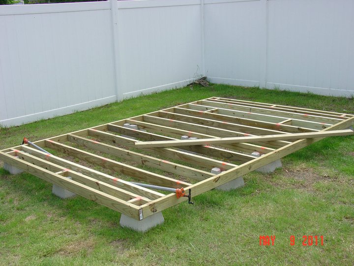 Wooden deck framing laid out on concrete blocks in a grassy yard with a white fence in the background