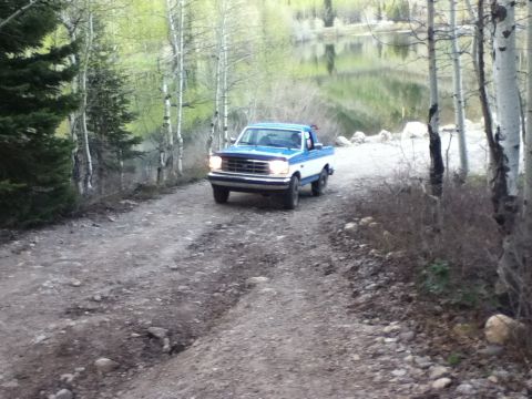 1994 Ford Bronco - Pickup truck navigates a rough dirt road near a lake, surrounded by trees.