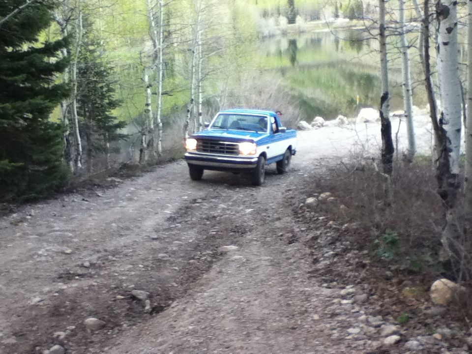 Blue and white pickup truck driving on a dirt road with trees and a lake in the background