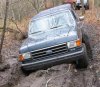 1989 Ford Bronco - A very small, low-resolution snapshot of a dark blue Ford Bronco nosed down over a muddy rutted bank on a wooded trail, front wheels flexed…