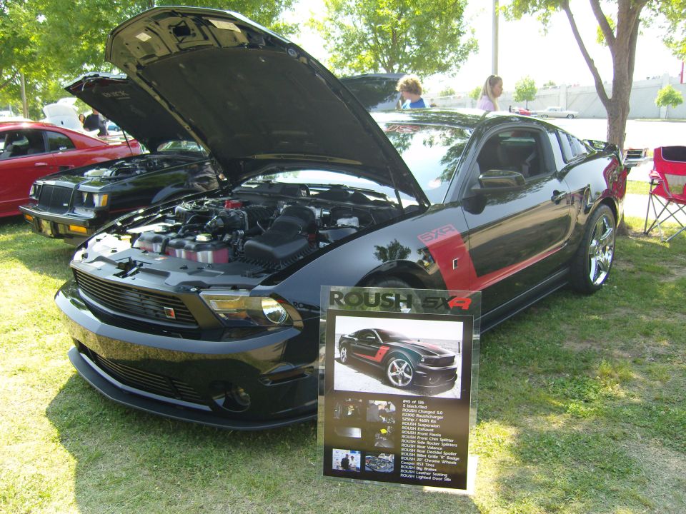 Black 2007 Ford Mustang Roush SX4 with hood open, parked on grass next to a display board. Another car is visible behind it