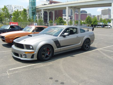 2007 Ford Mustang - A silver 2007 Ford Mustang with racing stripes and a large rear spoiler is parked in a lot with other cars.