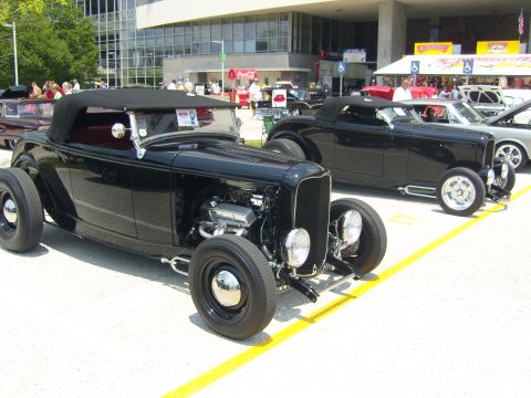 2007 Ford Mustang - Two black hot rod roadsters are parked side-by-side at an outdoor car show.