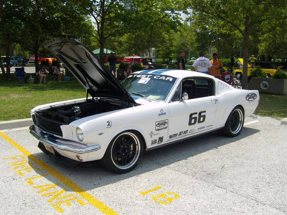 White 1965 Ford Mustang Fastback with blue racing stripes and number 66 on the door, hood open, parked on asphalt