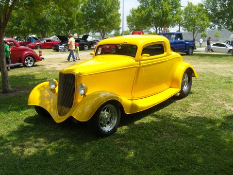 2007 Ford Mustang - A yellow 1934 Ford hot rod coupe is displayed at an outdoor car show with other vehicles present.