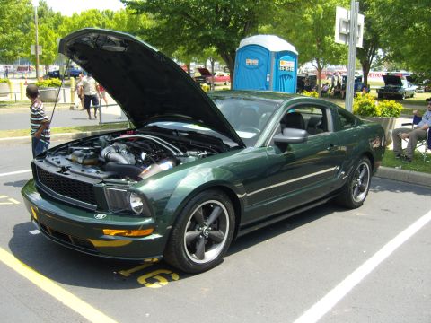 2007 Ford Mustang - A green Ford Mustang is parked with its hood up, revealing the engine compartment. A portable toilet is visible behind the car.