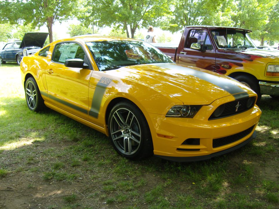 Yellow Ford Mustang Boss 302 with black stripe on hood and side. Parked next to a pickup truck