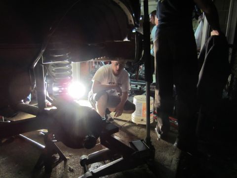 1992 Ford Bronco - Mechanics work on the rear suspension of a vehicle, illuminated by a bright work light.