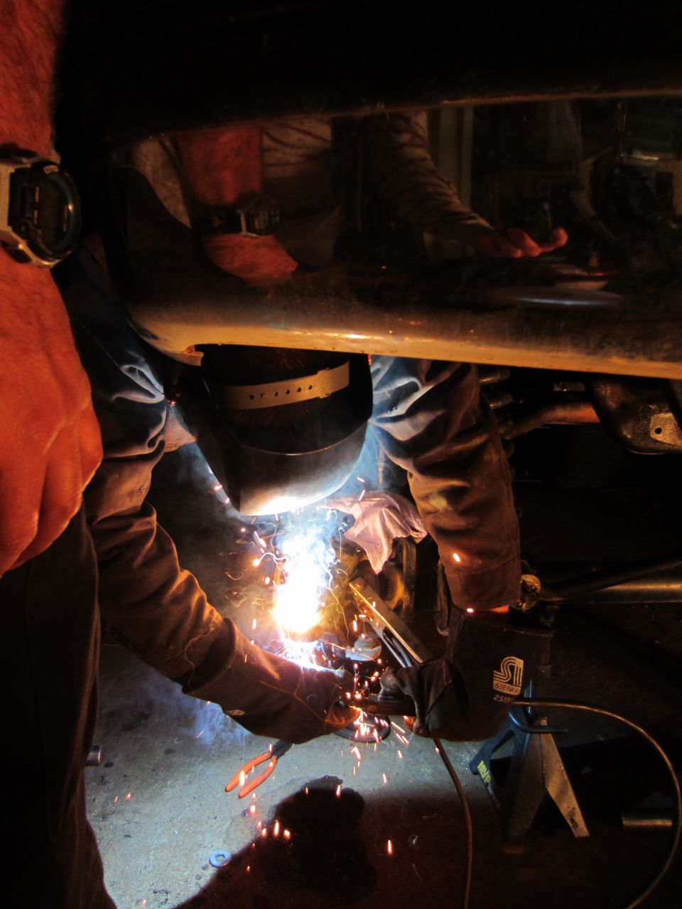 Welder in helmet sparks metal under vehicle frame, with jack stand visible
