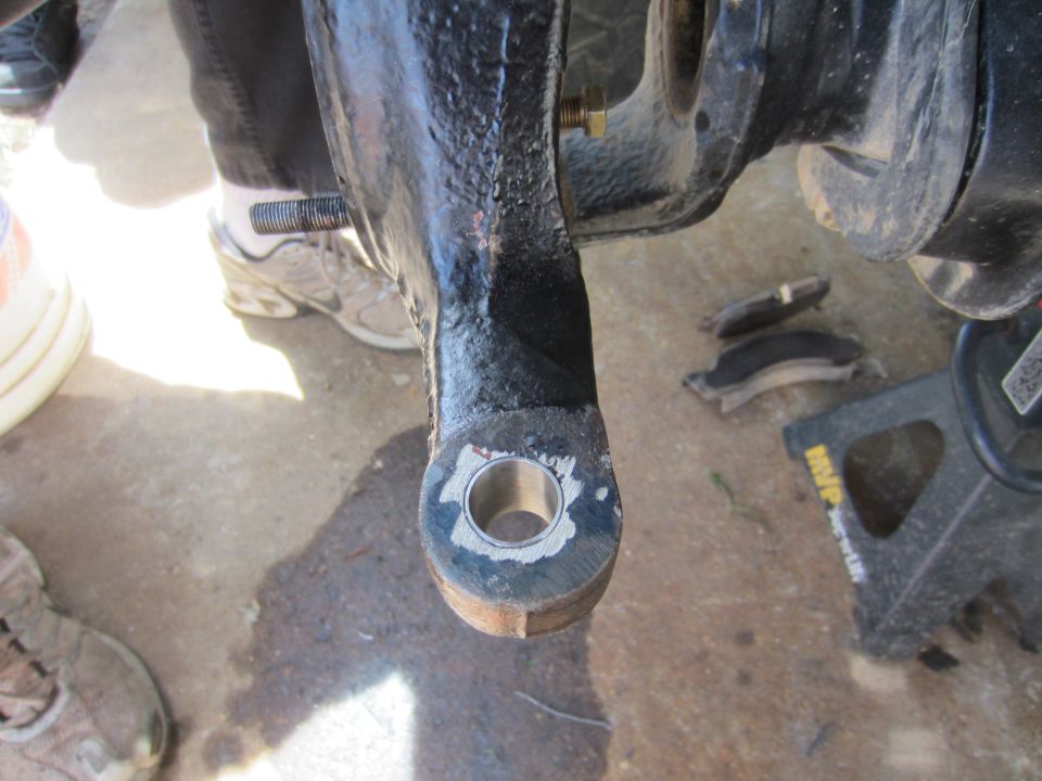 close-up of a vehicle's steering knuckle with a bolt and hole, brake pads and jack stand in background