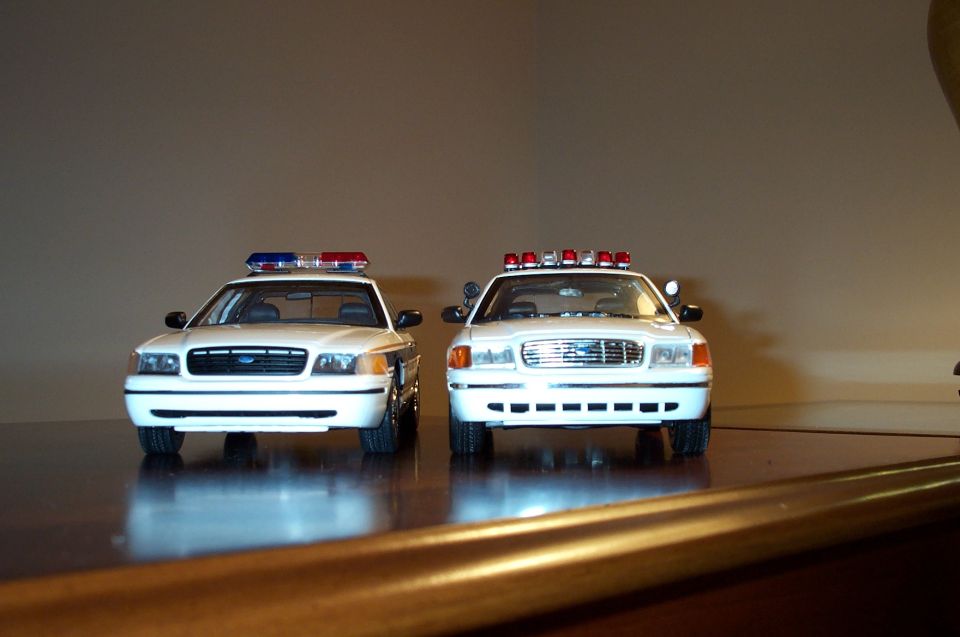 Two white Crown Victoria police model cars front view with roof light bars on wood shelf
