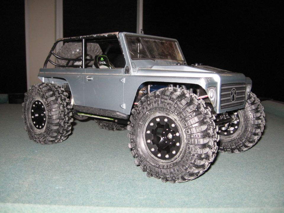 Silver RC rock crawler with large off-road tires and roll cage, viewed from the front three-quarter angle
