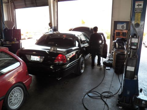 2003 Ford Crown Victoria - Rear view of a black sedan with California plate inside a service garage, technician working at the open driver door