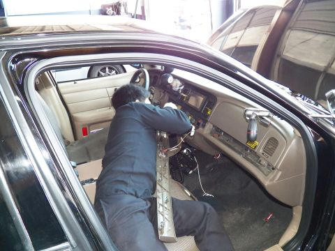 2003 Ford Crown Victoria - Man leans over the center console wiring a black sedan with tan trim, yellow airbag warning label visible.