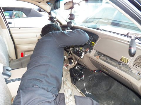 2003 Ford Crown Victoria - Worker reaches across tan dash with exposed wiring and a small screen, airbag warning label at right.