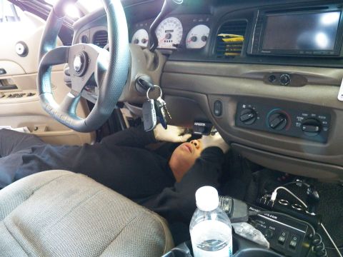 2003 Ford Crown Victoria - Technician works on his back beneath an aftermarket steering wheel and white gauges.