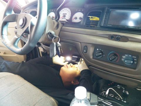 2003 Ford Crown Victoria - Man lies under the steering column with a flashlight, white-face gauges above him.