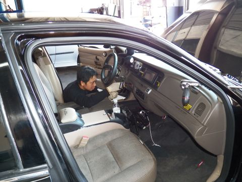 2003 Ford Crown Victoria - Worker sits in the driver seat of a black sedan, tan dash and center screen visible.