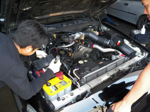 2003 Ford Crown Victoria - One technician wires the engine bay with a yellow battery while a second looks on.