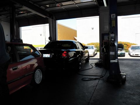2003 Ford Crown Victoria - Rear view of dark sedan in a repair-shop bay with red car at left and vehicles visible in the lot outside