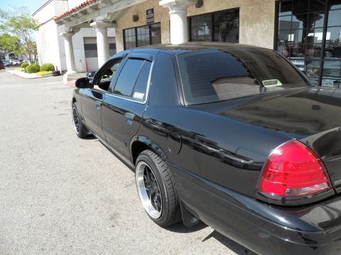 2003 Ford Crown Victoria - Glossy black full-size sedan shown from rear quarter with dark tint and aftermarket wheels at a shopping center