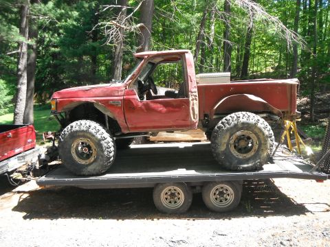 1988 Ford Bronco - Modified red pickup truck with oversized tires is transported on a flatbed trailer.