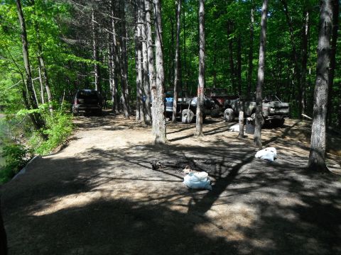 1988 Ford Bronco - Multiple off-road vehicles are parked on a dirt path surrounded by trees, with bags of trash visible on the ground.