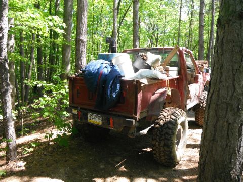 1988 Ford Bronco - A red pickup truck bed is filled with various items, including a blue jacket and white buckets, parked in a wooded area.