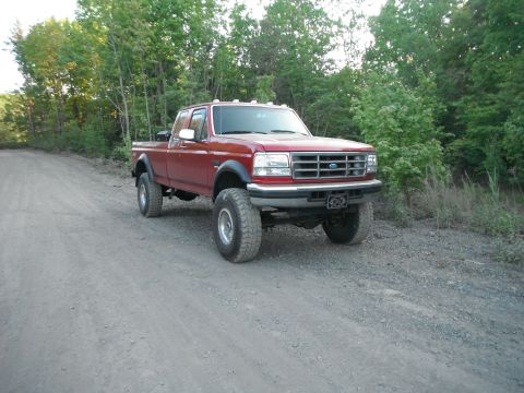 1988 Ford Bronco - A lifted red Ford F-Series pickup truck is parked on a dirt road surrounded by trees.