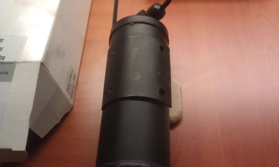 Black cylindrical automotive part with a metal band and visible mounting holes, next to a cardboard box